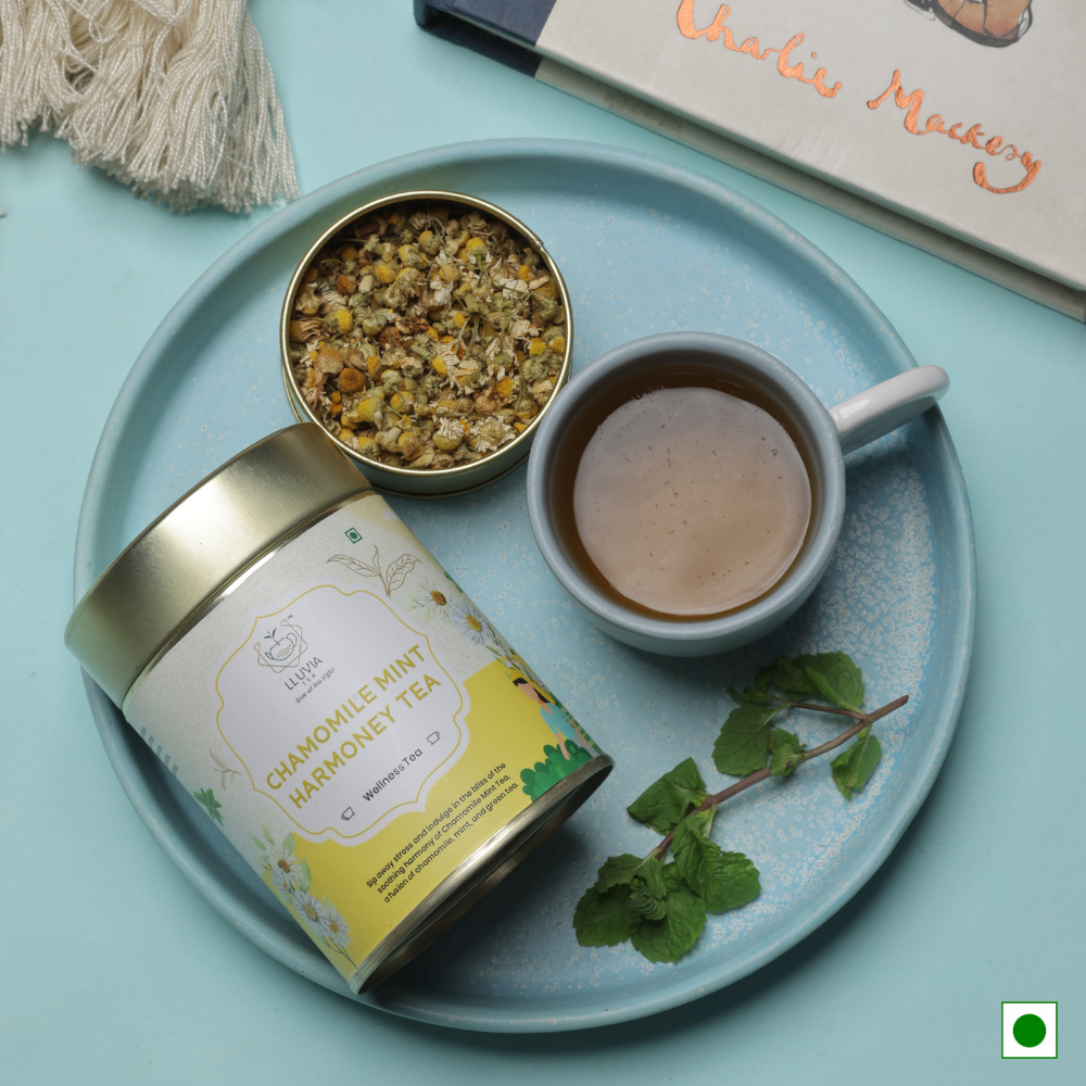 Steaming cup of Lluvia Chamomile Mint Green Tea with chamomile flowers, fresh mint leaves, loose green tea, and lemon slices on a rustic wooden table