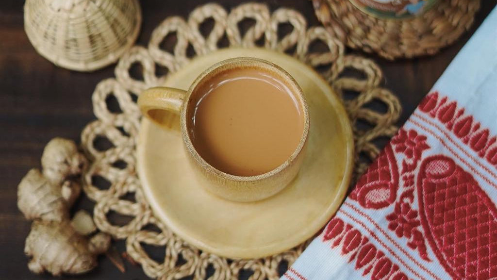Cup of tea on a saucer with a jar of kadak masala, ginger root, and a plant in the background.